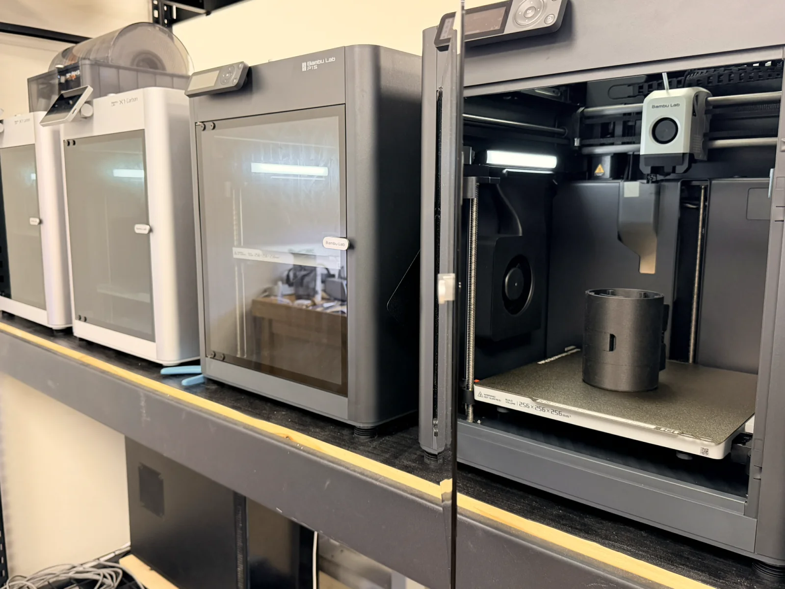 A row of 3D printers in the engineering space, one open with a printed part on its build plate.